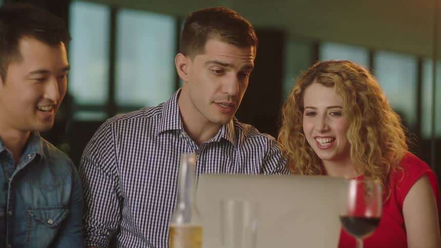 A group of people talk and laugh while looking at a laptop together at a table