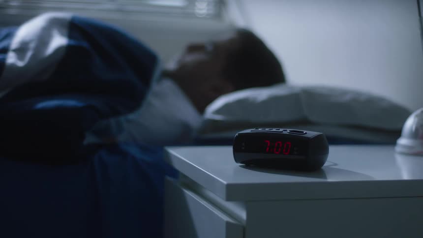 White man sleeping and then the wakes up to the digital alarm at 7am