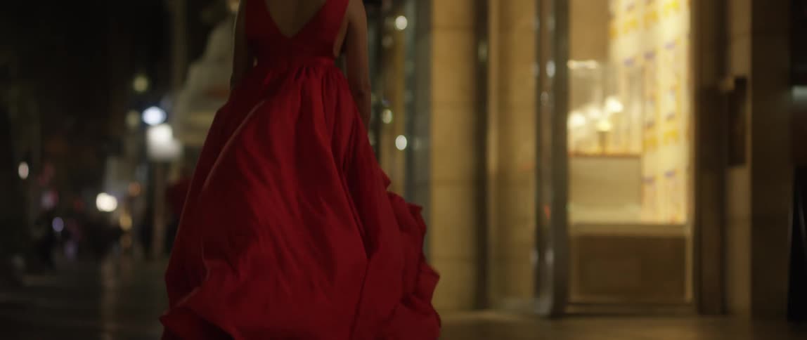 A woman with a red dress walking on a city street with buildings people and the sky in the background