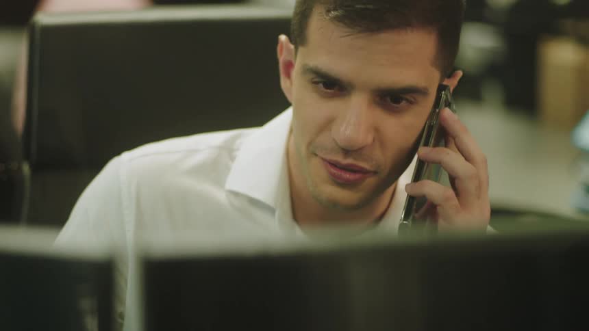Man smiles and talks on a smart phone while sitting in front of a computer monitor