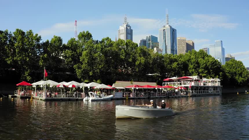 A motorboat travels past people sitting under umbrellas at an on the water restaurant in a park with city skyline behind