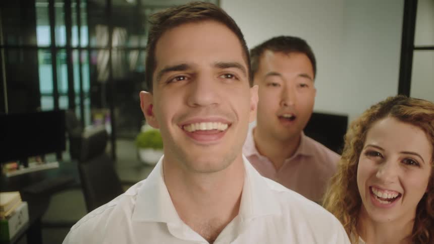 A man in an office pats another man on the back and a woman smiles and shakes her head