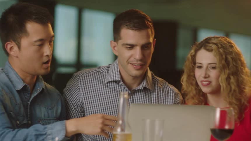Two men and a woman in a room at a table with a laptop beer bottle and glasses