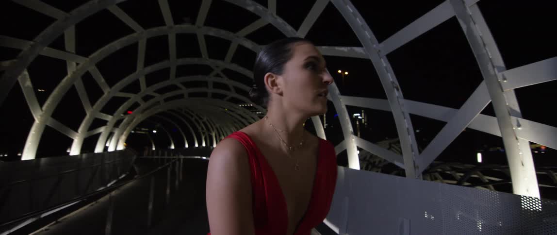 A woman with a dress and a necklace walking on a overpass with hand railing with buildings and the sky in the background