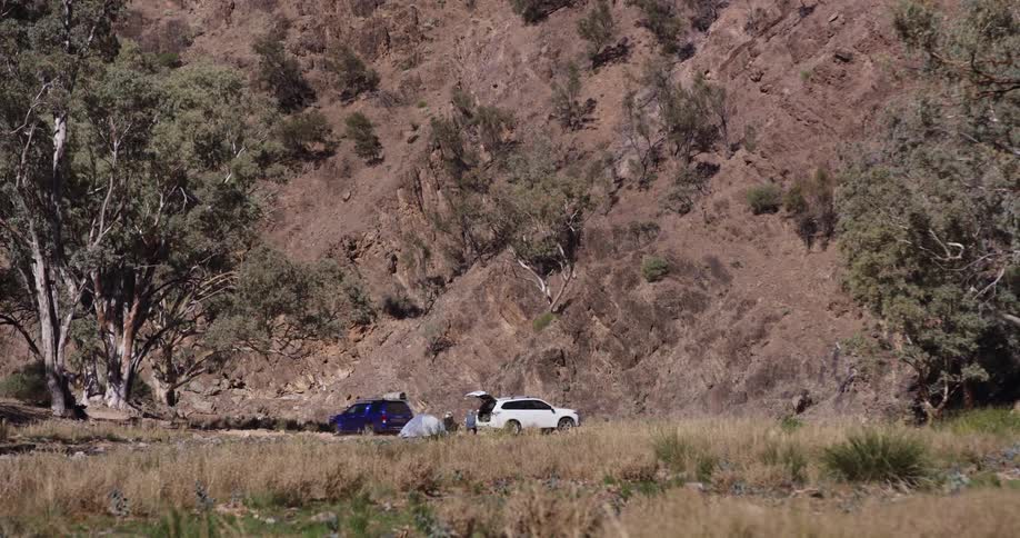 Two people sit by a tent behind their car as another car drives through water and water sprays up