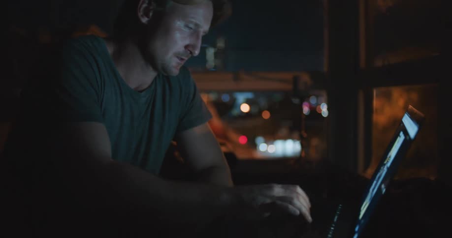 White man typing on his laptop alone at a dark room next to a big window