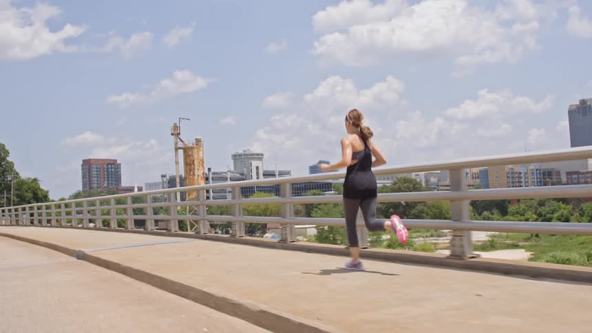 A woman jogs across a bridge with city buildings in the background