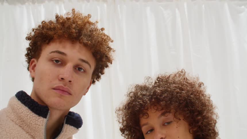 Curly man and woman stare down at the camera in front of white curtains