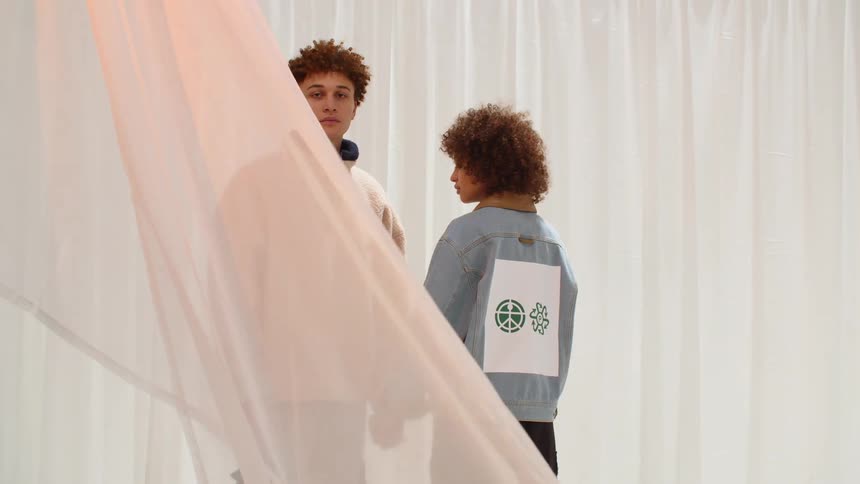 A man looks into the camera while standing motionlessly behind a waving curtain while holding the hand of a woman facing the opposite direction