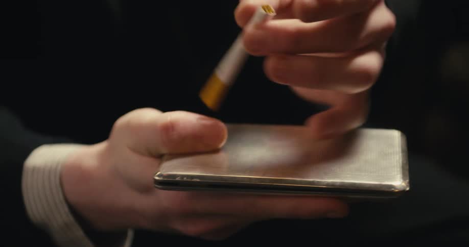 A man removes a cigarette from a metal case and taps the cigarette on it after closing