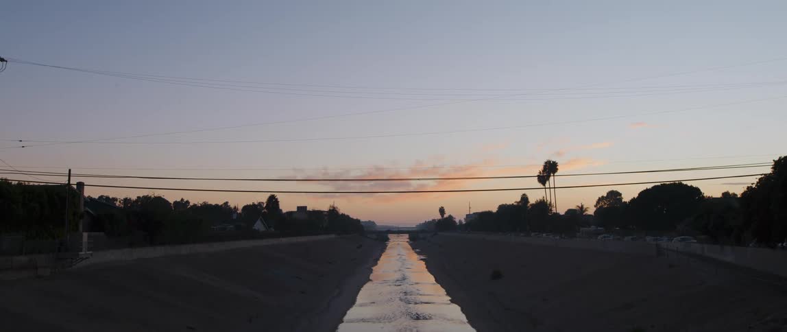 A canal with water with trees an overpass vehicles the sky and clouds in the background