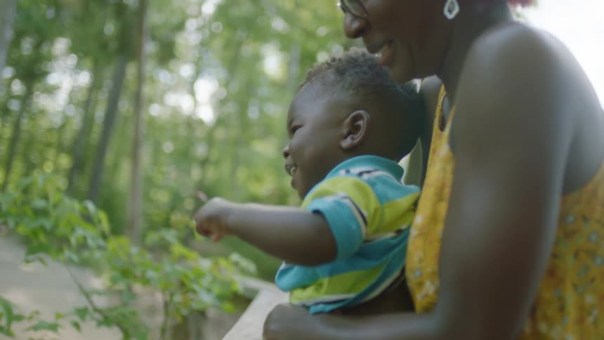 A young boy laughs as his mom holds him next to a ledge near a forest in a public park