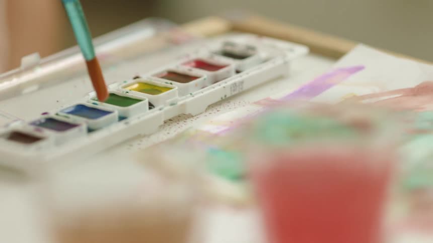 A person dips a paint brush in to a paint tray and then a glass of water