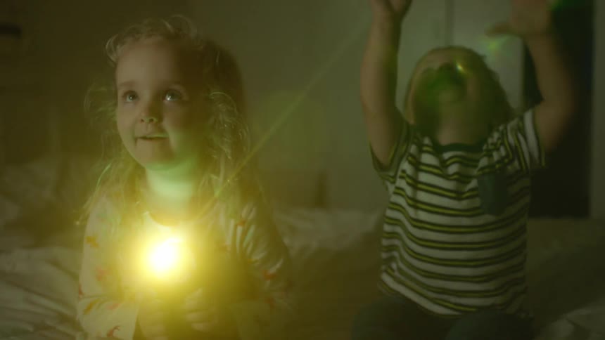 A toddler raises his arms and laughs as his sister shines a flashlight on the ceiling of a bedroom as they sit side by side on a bed