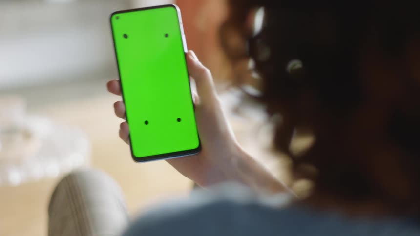 A natural lifestyle shot of a woman sitting indoors as she raises her smartphone with a green screen and tracking markers from her side to look at the display before lowering it again. Filmed in soft daylight with a cozy, realistic atmosphere.