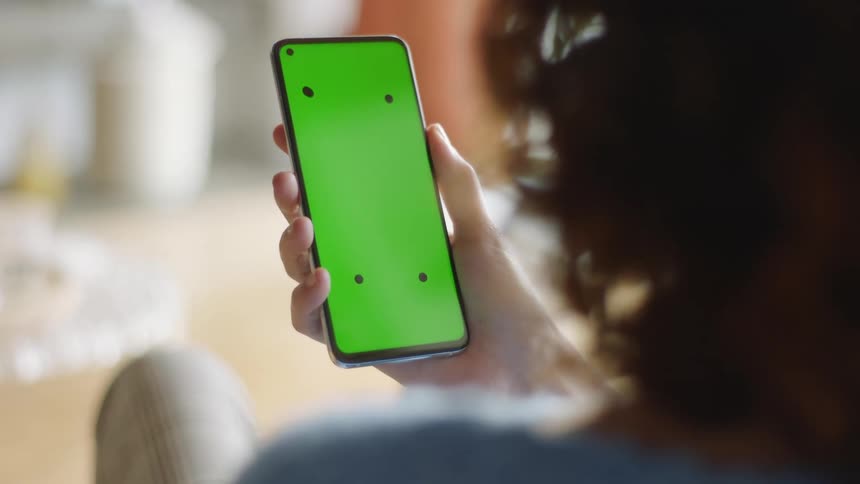 Natural lifestyle shot of a woman sitting indoors raising a smartphone with green screen and tracking marks to look at the display, then lowering it. Filmed in soft daylight, perfect for VFX, app mockups, and digital tech projects.