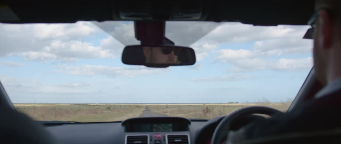 A man wearing sunglasses drives on an empty desert road on a blue sky day