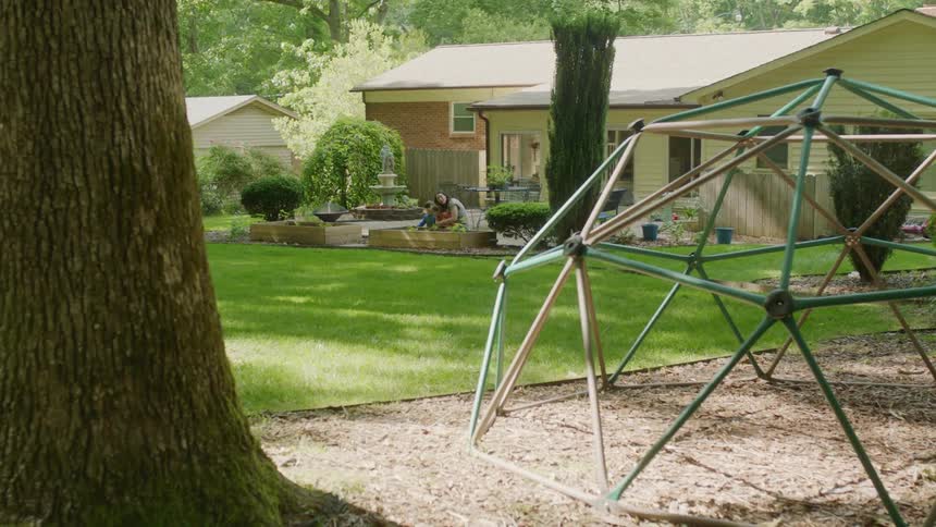 A jungle gym next to a tree with a woman two children and a house in the background