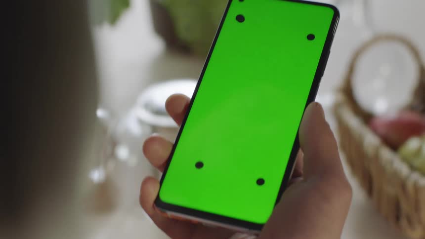 Close-up of hand holding smartphone with green screen and tracking marks as camera tilts upward following movement before device exits frame. Filmed indoors in soft natural light, perfect for VFX, app mockups, and digital tech projects.