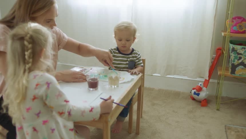 A mom kneels beside a small table and helps her two children paint