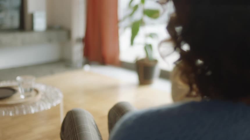 A natural lifestyle shot of a woman sitting in a bright living room as she raises a smartphone with a green screen and tracking markers to look at the display before lowering it again. Filmed in soft daylight.