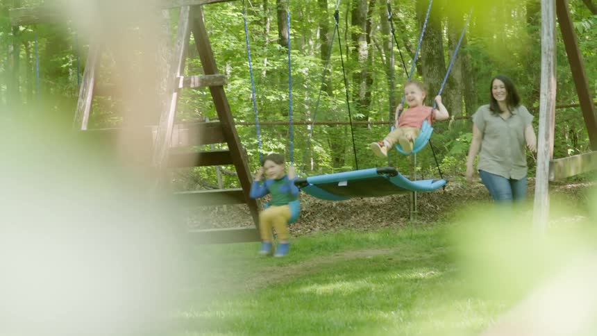 A mom walks back and forth pushing her two kids on the back yard swings