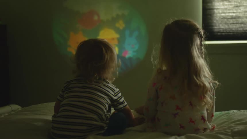 A brother and sister sit on a bed watching an image projected on the wall