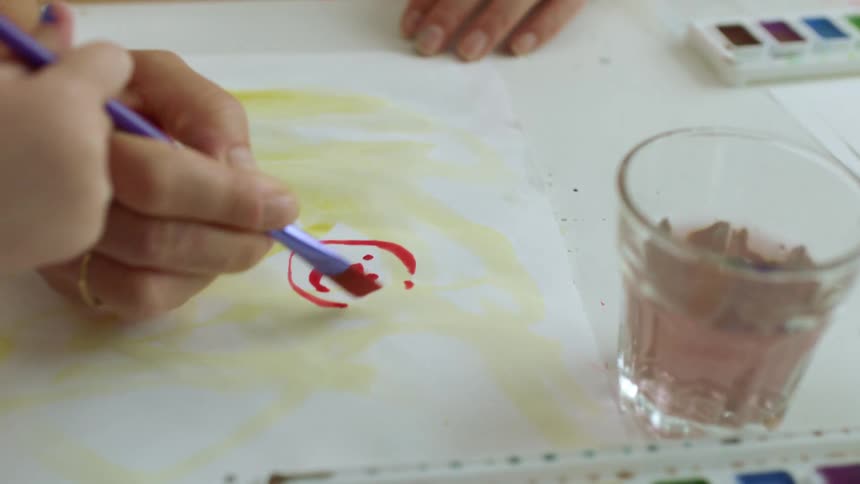 A child holds the paint brush as mom draws with a brush and watercolors