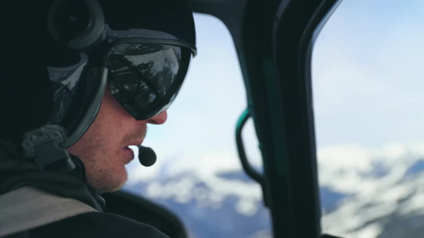 A man with a headset and a helmet in the cockpit of an aircraft with a mountain range the sky and clouds in the background