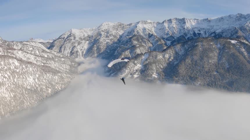 A paraglider glides over a foggy mountain valley between snow covered forested mountains on a sunny day