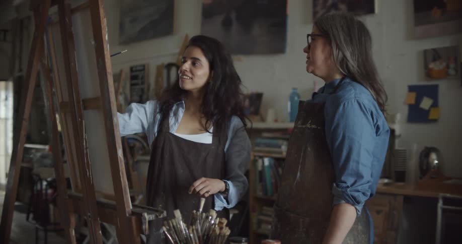 A young woman paints on a canvas while be instructed by an older woman in a workshop during the day