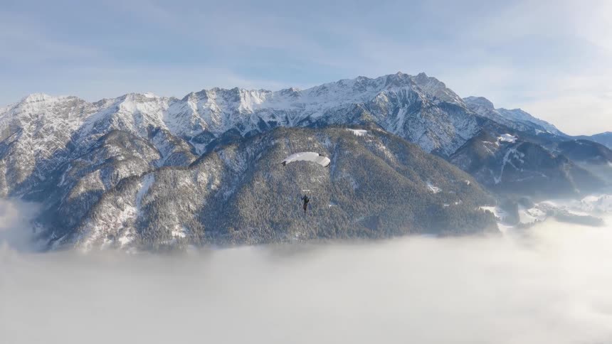 Skydiver wearing a parachute is flying high over clouds near snow covered mountains