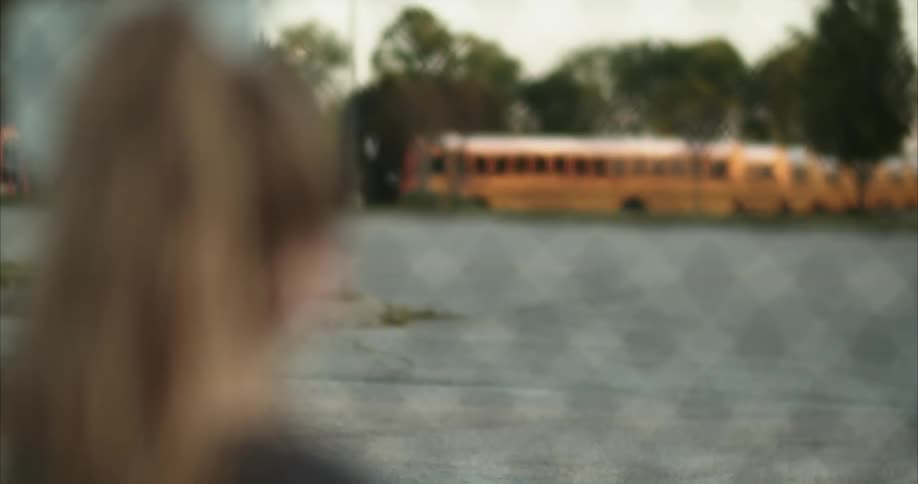 A young girl stands at a fence and looks at the school buses across the lot