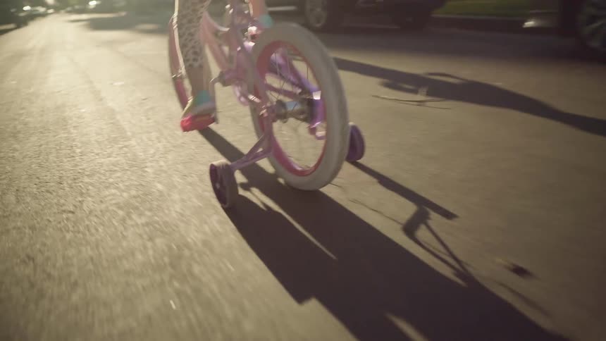 A girl and boy ride bicycles with training wheels down a neighborhood street during the day