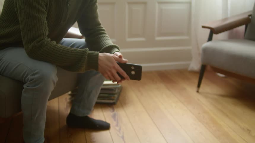 A person in a room sitting on a couch holding a smart phone with a wall and a stack of books in the background