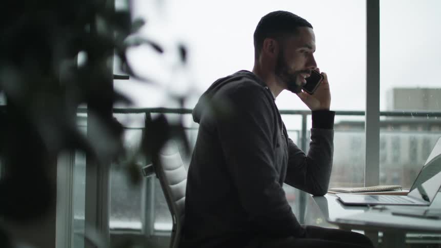 A man is talking on his cell phone at his desk by the window