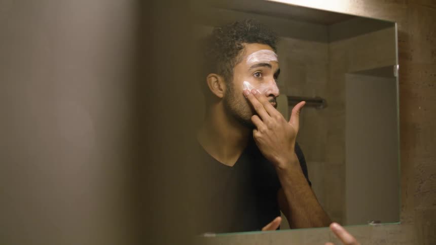 A man in a bathroom in front of a mirror applying cream to his face