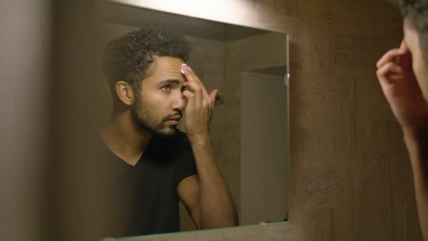 A man standing before a mirror in a bathroom spreads cosmetic cream across his face