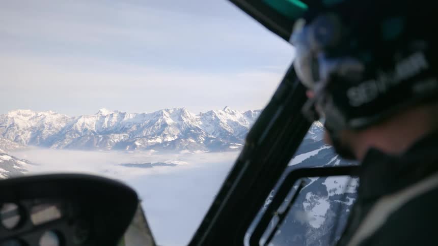 A pilot in the front of a moving helicopter looking down over a snow covered mountain range