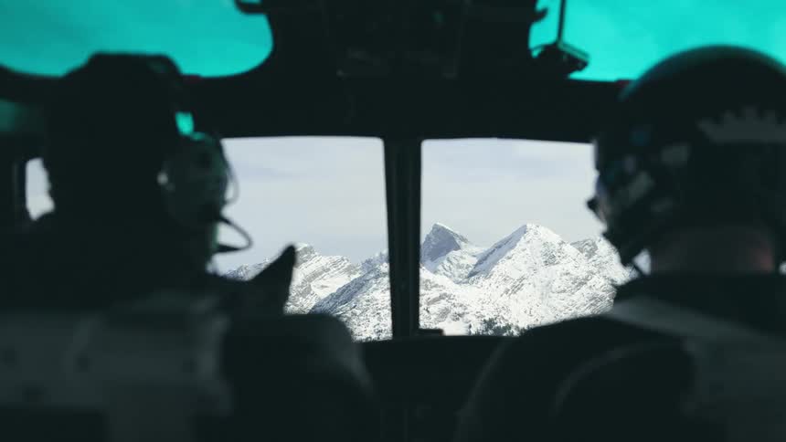 Two people sitting on the pilot seats of a helicopter flying over the snow covered mountain peaks