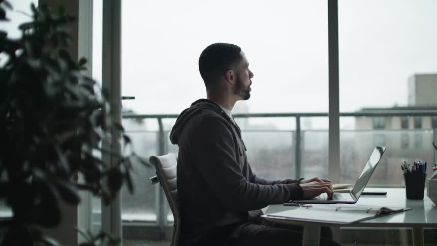 A man is typing on a laptop while sitting next to a window