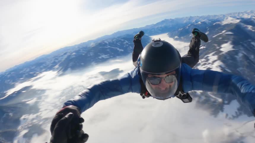 A man skydiving over the snow covered peaks of a mountain range hold hands with the cameraman