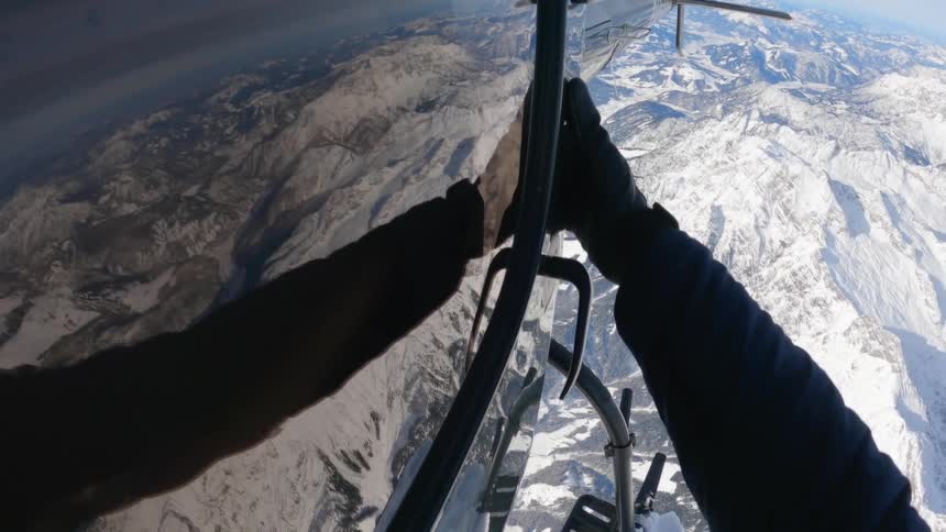A person with gloves and a coat standing outside a helicopter in the air with a mountain range the sky and clouds in the background
