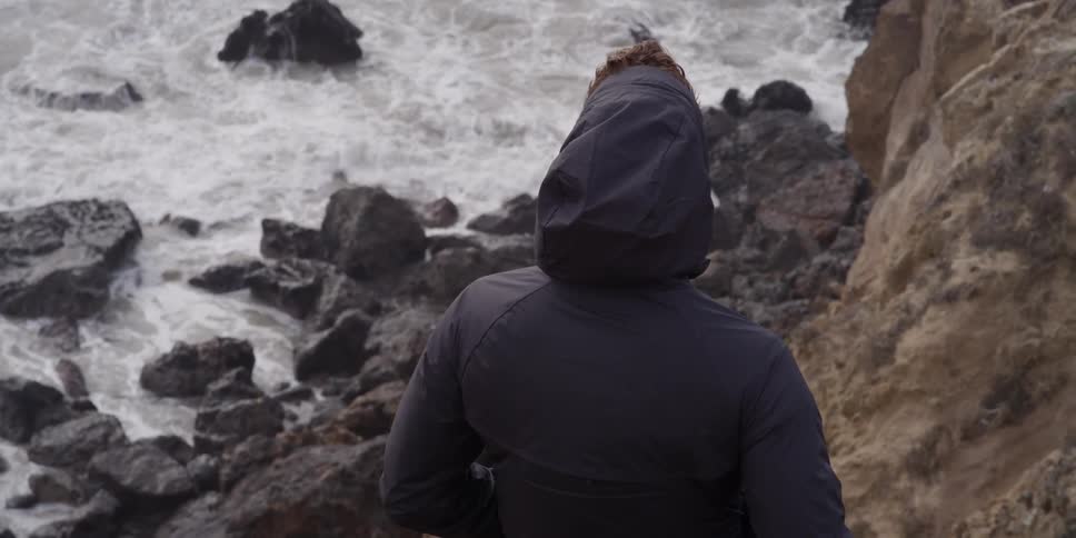 Hooded person looking down at waters crashing upon a rocky area