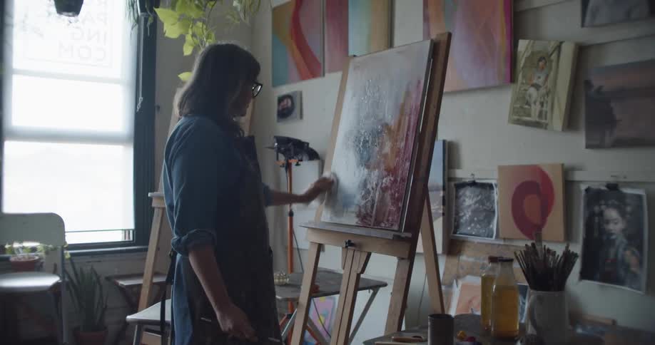 Woman wearing an apron painting with a small brush over canvas on an easel inside a studio