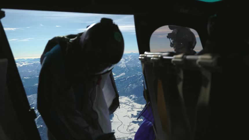 A man wearing paragliding gear stands at the entrance of a helicopter in the air over a snowy mountain range