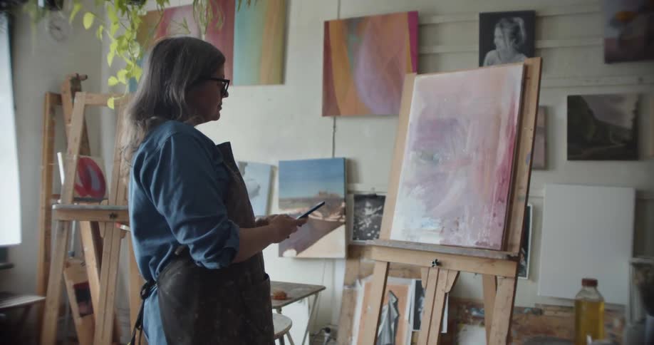 A woman in a room with glasses and an apron taking a picture of a canvas with a smart phone