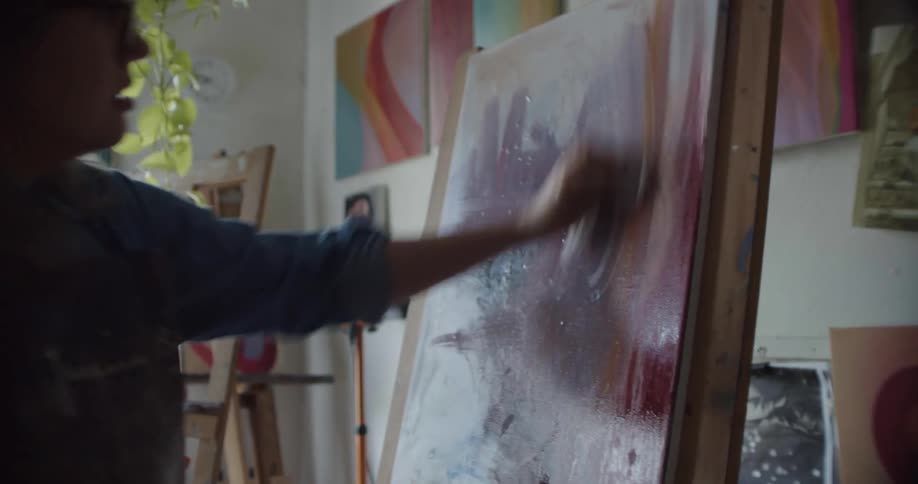 A woman wearing glasses applies paint to a canvas on a standing easel