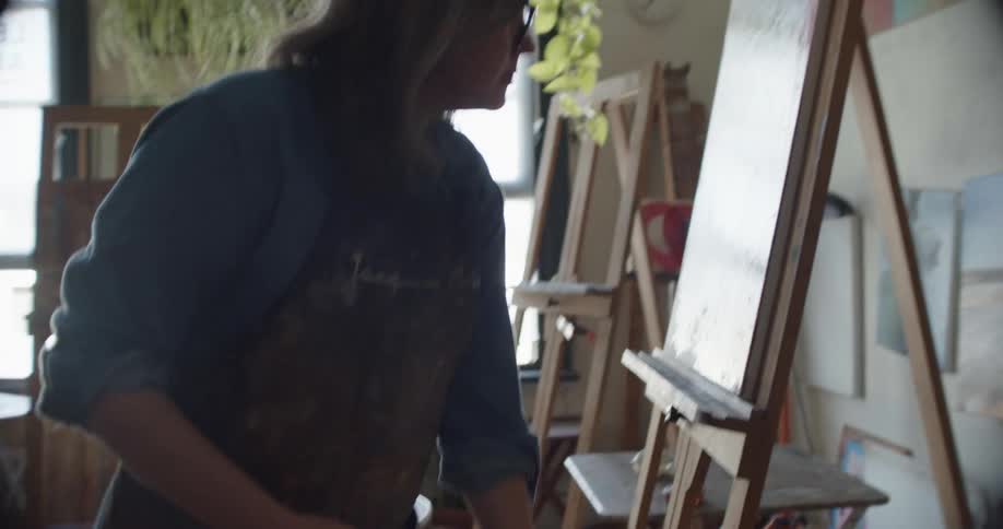 A woman stands before a canvas atop an easel and applies paint with a brush while pausing to plan her next steps