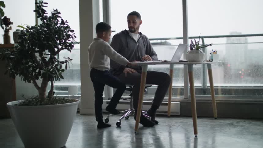 A man sitting in a chair at a desk typing on a laptop as a boy walks up to him
